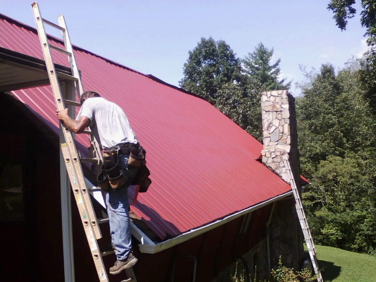 Roofer on a ladder beside a red metal roof and stone chimney — Soffit & Fascia Repair by Roofing Brothers in Maria Stein